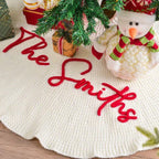 Cream knitted tree skirt with The Smiths embroidered in red, beside a snowman and presents