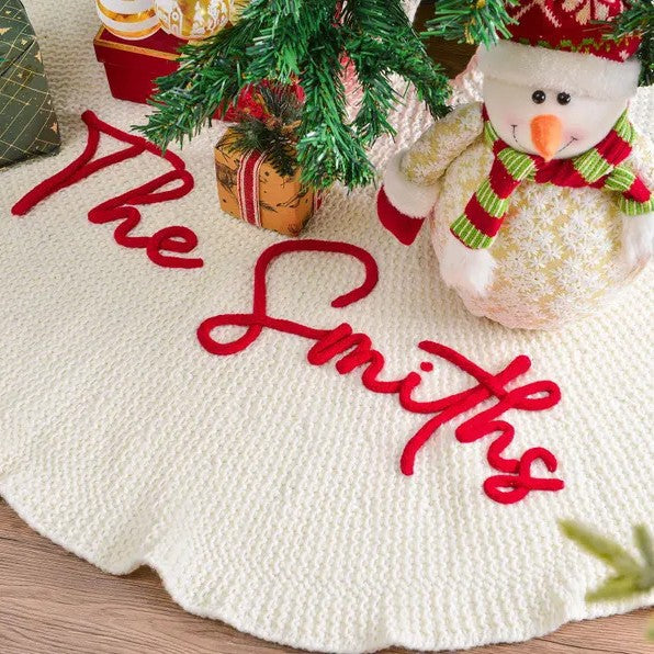 Cream knitted tree skirt with The Smiths embroidered in red, beside a snowman and presents