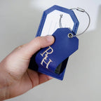 Blue suitcase tag with RH initials stitched in gold, held in one hand to show its size